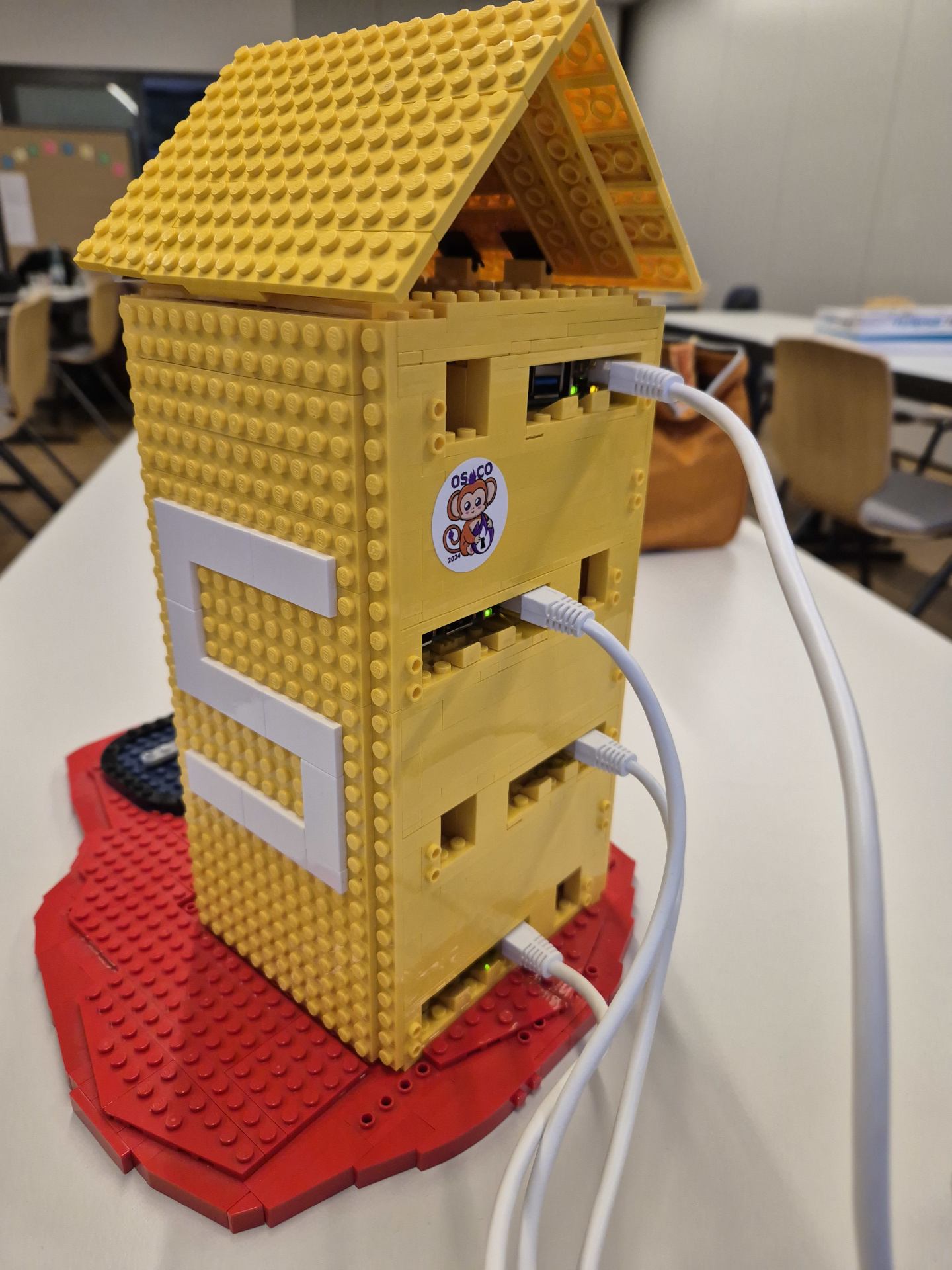 The picture shows a cluster made of multiple Raspberry Pies. The enclosure is the juice package from the logo of the OWASP juice shop and is made out of lego bricks. Someone put a osco 2024 sticker on it.