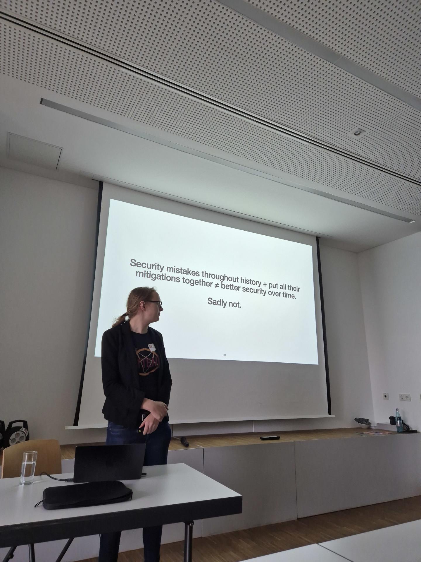 The picture shows aa scene from the keynote 'History repeating itself'. Bianca, the speaker, is talking in the front. In the background is one page of the presentation on the screen. It reads 'Security mistakes throughout history + put all their mitigations together ≠ better security over time. Sadly not'.